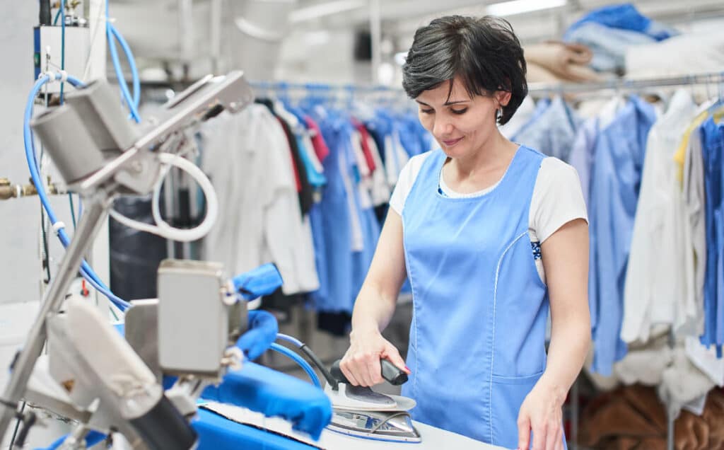 A woman in a blue uniform operates an ironing machine in a laundry or dry-cleaning facility. She is focused on her task, with racks of clothing visible in the background. The scene highlights a bright and professional work environment where competitive prices meet exceptional service.